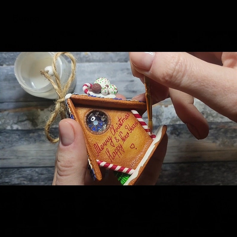 May include: A gingerbread house ornament with a red and white candy cane, green frosting, and a blue and white window. The ornament is decorated with the text "Merry Christmas & Happy New Year".