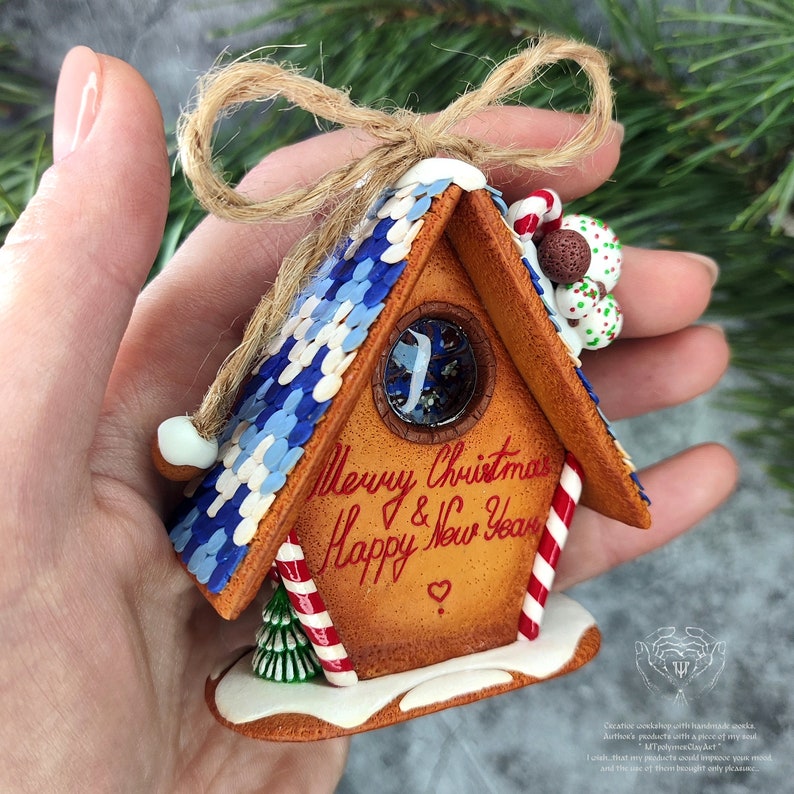 May include: A handmade gingerbread house ornament with a blue and white roof, candy cane accents, and a window with glitter. The ornament is decorated with the text "Merry Christmas & Happy New Year" in red.
