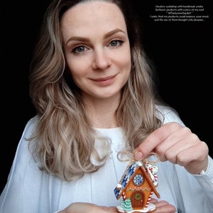 May include: A gingerbread house ornament with blue and white icing, candy canes, and a green tree. The ornament is held in the hands of a person.