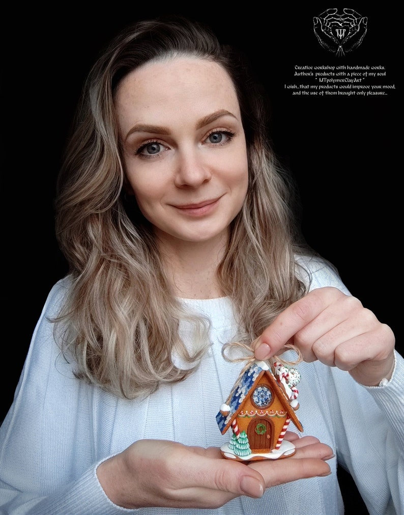 May include: A gingerbread house ornament with blue and white icing, candy canes, and a green tree. The ornament is held in the hands of a person.
