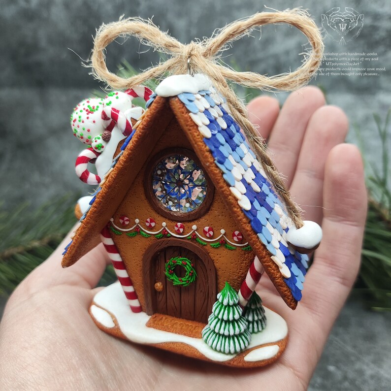 May include: A gingerbread house Christmas ornament with a blue and white roof, candy cane accents, a wreath on the door, and a small evergreen tree in front.