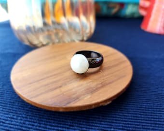 May include: A black ring with a white pearl set in the centre. The ring is on a wooden surface.