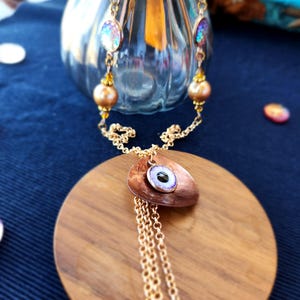 Puede incluir: Un collar de cadena dorada con un colgante de cobre con un diseño de ojo morado y azul. El collar se muestra sobre una superficie de madera.