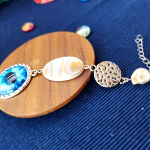 Puede incluir: Un brazalete de plata con un dije de ojo azul, un dije de concha blanca, un dije de plata con un diseño texturizado y un dije de concha blanca. El brazalete está sobre una superficie de madera.