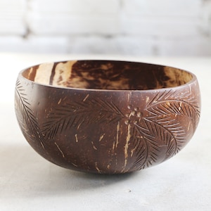 May include: A dark brown coconut bowl with carved palm leaf designs. The bowl has a smooth, rounded shape and a natural, textured interior. The exterior features intricate carvings of palm leaves and small floral accents.