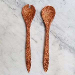 Könnte beinhalten: Ein Set aus zwei Salatservierern aus Kokosnussholz. Die Servierer haben eine natürliche braune Farbe und eine strukturierte Oberfläche. Der Servierer links hat ein kleines Loch oben zum Aufhängen.