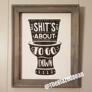 May include: Framed bathroom art with a humorous message. The black and white print features a toilet graphic with the phrase "Shit's About To Go Down." The wooden frame has a gray finish.