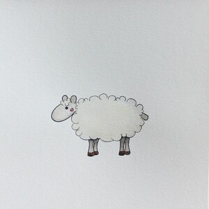 Peut inclure: Une carte blanche illustrant un mouton blanc avec des yeux et des sabots noirs. La carte est suspendue à un clip en bois.
