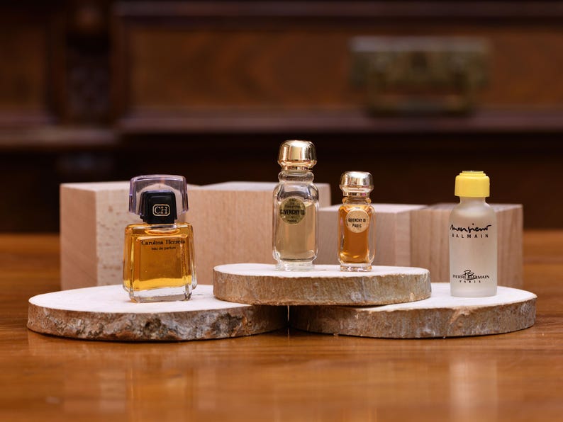 K&ouml;nnte beinhalten: Vier Miniatur-Parf&uuml;mflaschen in verschiedenen Formen und Gr&ouml;&szlig;en werden auf Holzplattformen pr&auml;sentiert. Die Flaschen sind mit bernsteinfarbenen Fl&uuml;ssigkeiten gef&uuml;llt und haben goldene oder transparente Kappen. Der Hintergrund umfasst Holzbl&ouml;cke und eine dunkle Holzoberfl&auml;che.