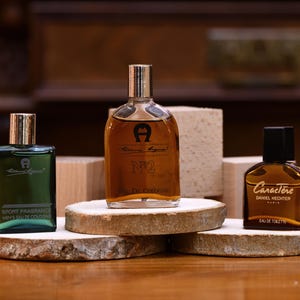 K&ouml;nnte beinhalten: Drei Parf&uuml;mflaschen auf Holzuntersetzern. Die Flaschen haben silberne und schwarze Verschl&uuml;sse und sind in verschiedenen Formen und Gr&ouml;&szlig;en erh&auml;ltlich. Die Etiketten tragen die Aufschrift "Aigner", "N2" und "Caract&egrave;re Daniel Hechter". Die Flaschen stehen auf einer Holzoberfl&auml;che.