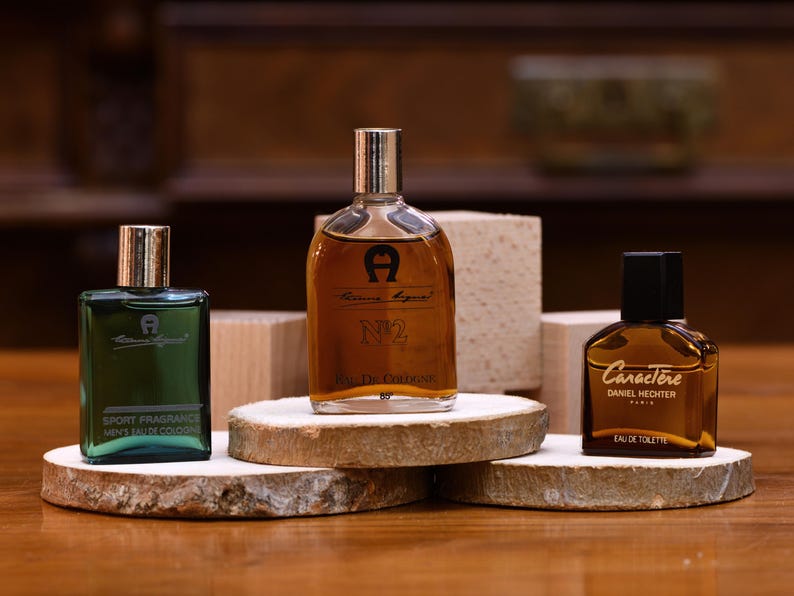 K&ouml;nnte beinhalten: Drei Parf&uuml;mflaschen auf Holzuntersetzern. Die Flaschen haben silberne und schwarze Verschl&uuml;sse und sind in verschiedenen Formen und Gr&ouml;&szlig;en erh&auml;ltlich. Die Etiketten tragen die Aufschrift "Aigner", "N2" und "Caract&egrave;re Daniel Hechter". Die Flaschen stehen auf einer Holzoberfl&auml;che.