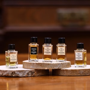 K&ouml;nnte beinhalten: F&uuml;nf Miniatur-Chanel-Parf&uuml;mflaschen mit schwarzen Kappen und Etiketten, pr&auml;sentiert auf kleinen Holzplattformen. Die Flaschen enthalten eine hellbernsteinfarbene Fl&uuml;ssigkeit und sind mit der Marke Chanel und den Produktnamen versehen.