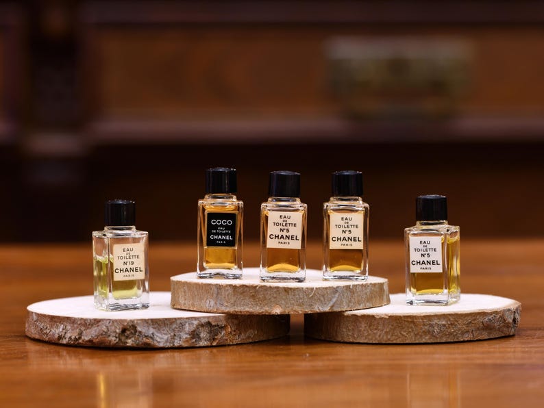 K&ouml;nnte beinhalten: F&uuml;nf Miniatur-Chanel-Parf&uuml;mflaschen mit schwarzen Kappen und Etiketten, pr&auml;sentiert auf kleinen Holzplattformen. Die Flaschen enthalten eine hellbernsteinfarbene Fl&uuml;ssigkeit und sind mit der Marke Chanel und den Produktnamen versehen.