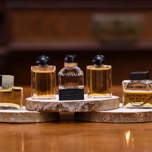 K&ouml;nnte beinhalten: F&uuml;nf Miniatur-Parf&uuml;mflaschen werden auf kleinen, runden Holzst&auml;ndern pr&auml;sentiert. Die Flaschen variieren in Form und Design, mit klarem Glas und dunklen Verschl&uuml;ssen. Die D&uuml;fte sind bernsteinfarben, und die Flaschen sind auf einer Holzoberfl&auml;che angeordnet.