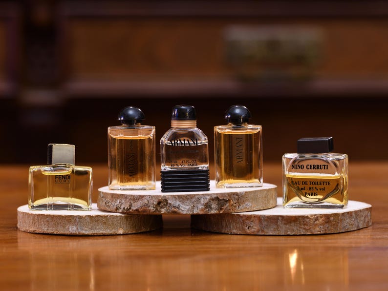 K&ouml;nnte beinhalten: F&uuml;nf Miniatur-Parf&uuml;mflaschen werden auf kleinen, runden Holzst&auml;ndern pr&auml;sentiert. Die Flaschen variieren in Form und Design, mit klarem Glas und dunklen Verschl&uuml;ssen. Die D&uuml;fte sind bernsteinfarben, und die Flaschen sind auf einer Holzoberfl&auml;che angeordnet.