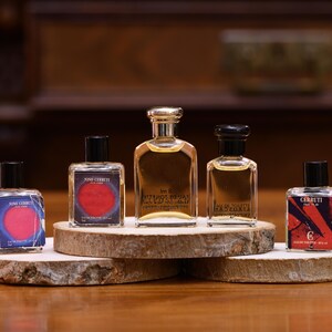 K&ouml;nnte beinhalten: F&uuml;nf Parf&uuml;mflaschen in verschiedenen Formen und Gr&ouml;&szlig;en werden auf einem Holzst&auml;nder pr&auml;sentiert. Die Flaschen haben schwarze Kappen und klares Glas, das die bernsteinfarbene Fl&uuml;ssigkeit zeigt. Die Etiketten zeigen Text und farbenfrohe Designs.