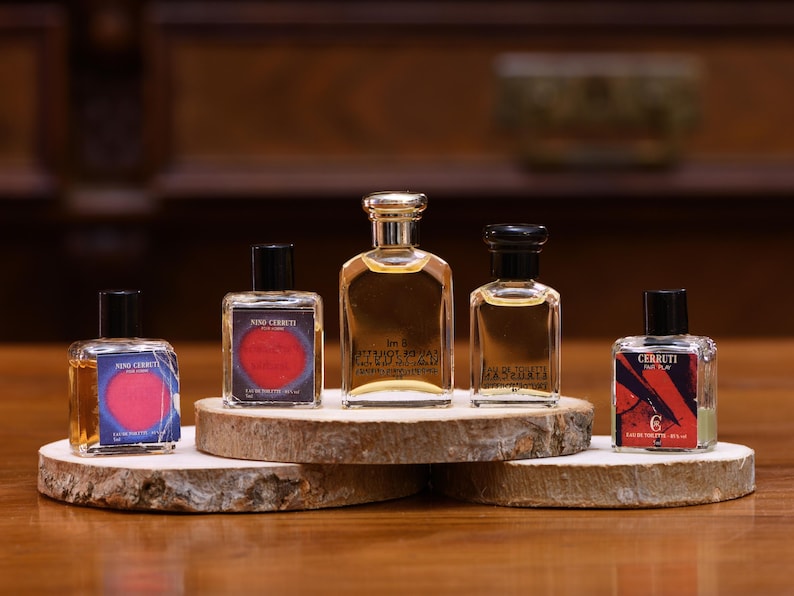 K&ouml;nnte beinhalten: F&uuml;nf Parf&uuml;mflaschen in verschiedenen Formen und Gr&ouml;&szlig;en werden auf einem Holzst&auml;nder pr&auml;sentiert. Die Flaschen haben schwarze Kappen und klares Glas, das die bernsteinfarbene Fl&uuml;ssigkeit zeigt. Die Etiketten zeigen Text und farbenfrohe Designs.