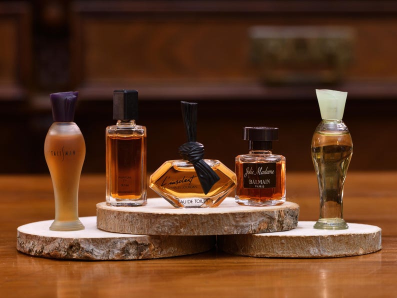 K&ouml;nnte beinhalten: F&uuml;nf Miniatur-Parf&uuml;mflaschen in verschiedenen Formen und Farben werden auf Holzscheiben pr&auml;sentiert. Die Flaschen haben unterschiedlich farbige Fl&uuml;ssigkeiten und Verschl&uuml;sse. Die Flaschen sind auf einer Holzoberfl&auml;che angeordnet.