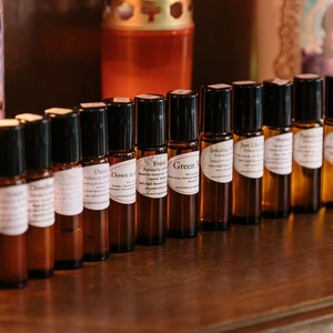 May include: A row of small brown glass bottles with black roll-on tops. Each bottle has a white label with a different name printed on it, such as "Fetish", "Green", "Inkblots", and "Just Like That".