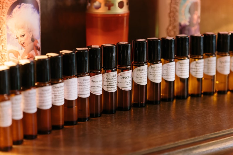 May include: A row of small brown glass bottles with black roll-on tops. Each bottle has a white label with a different name printed on it, such as "Fetish", "Green", "Inkblots", and "Just Like That".