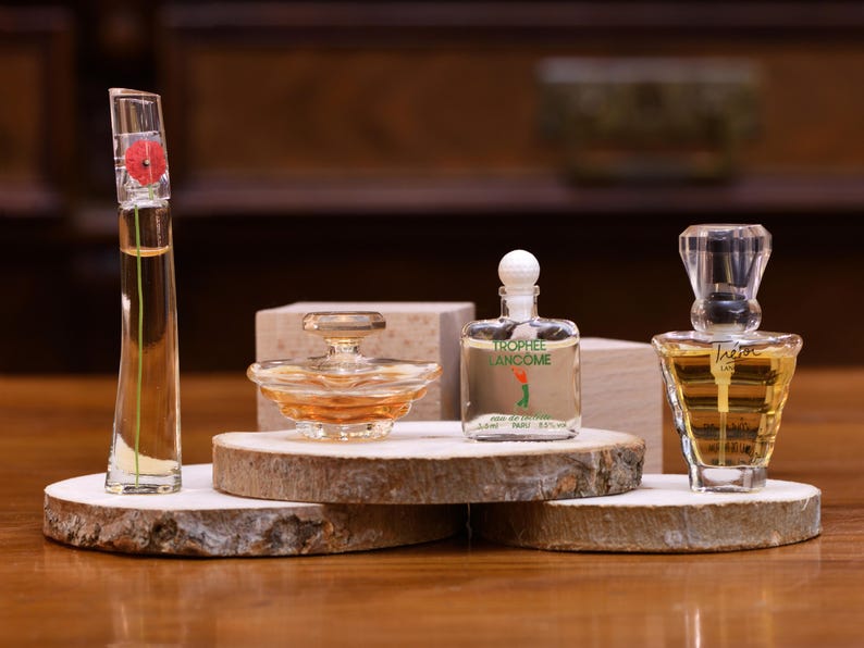K&ouml;nnte beinhalten: Vier Parf&uuml;mflaschen in verschiedenen Formen und Gr&ouml;&szlig;en werden auf Holzplattformen pr&auml;sentiert. Die Flaschen sind klar, mit bernsteinfarbenen und roten Fl&uuml;ssigkeiten. Eine Flasche hat eine rote Blume oben, und eine andere hat den Text "Trophe Lancome".