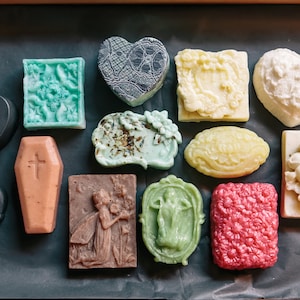 May include: Assortment of handmade soap bars in various shapes and designs, including a coffin, skull, heart, and floral patterns. The soaps are in shades of black, white, green, brown, and pink.