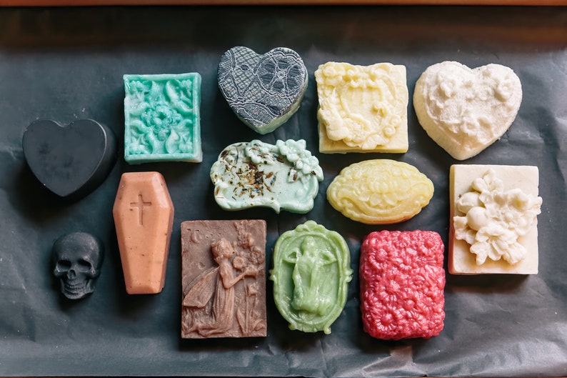 May include: Assortment of handmade soap bars in various shapes and designs, including a coffin, skull, heart, and floral patterns. The soaps are in shades of black, white, green, brown, and pink.