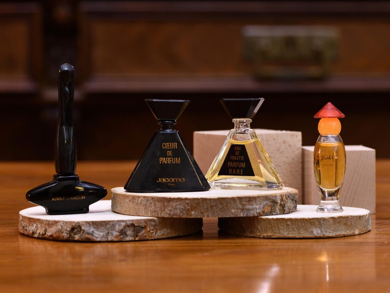 K&ouml;nnte beinhalten: Eine Sammlung von Vintage-Parf&uuml;mflaschen in verschiedenen Formen und Gr&ouml;&szlig;en. Die Flaschen werden auf kleinen Holzscheiben pr&auml;sentiert und umfassen eine schwarze Flasche mit einer einzigartigen Form, eine dreieckige schwarze Flasche mit dem Text "COEUR DE PARFUM" und eine klare Flasche mit dem Text "EAU DE TOILETTE PARFUM RARE".