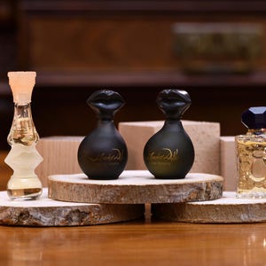 K&ouml;nnte beinhalten: Eine Auswahl an Parf&uuml;mflaschen, pr&auml;sentiert auf Holzscheiben. Die Flaschen variieren in Form und Farbe, darunter eine transparente, sanduhrf&ouml;rmige Flasche, zwei schwarze Flaschen mit lippenf&ouml;rmigen Verschl&uuml;ssen und eine rechteckige Flasche mit blauem Deckel.
