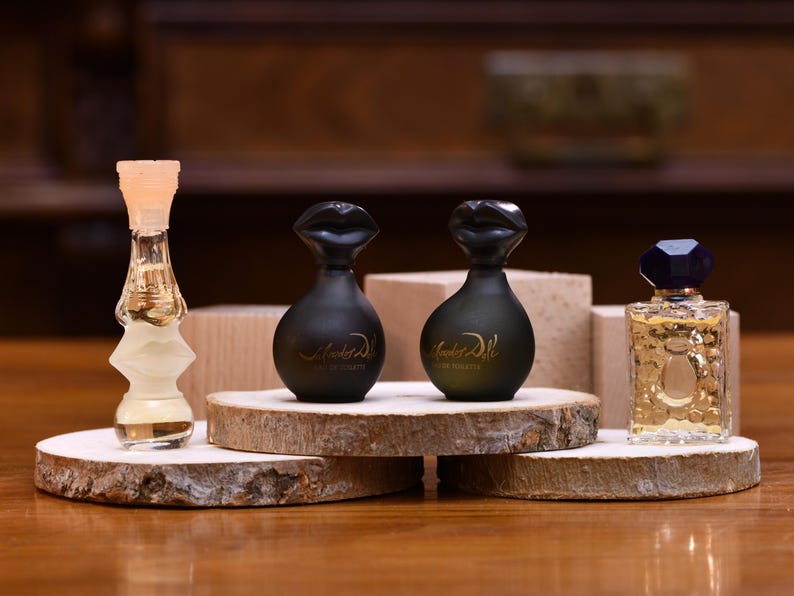 K&ouml;nnte beinhalten: Eine Auswahl an Parf&uuml;mflaschen, pr&auml;sentiert auf Holzscheiben. Die Flaschen variieren in Form und Farbe, darunter eine transparente, sanduhrf&ouml;rmige Flasche, zwei schwarze Flaschen mit lippenf&ouml;rmigen Verschl&uuml;ssen und eine rechteckige Flasche mit blauem Deckel.