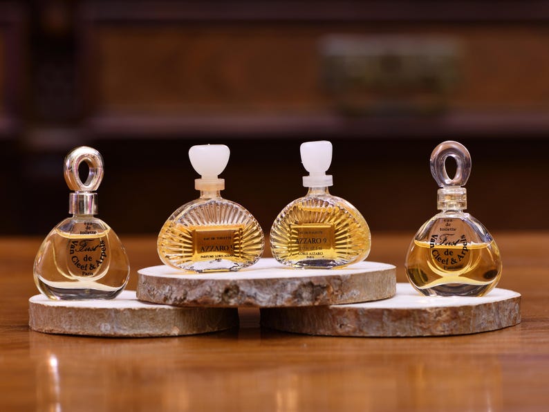 K&ouml;nnte beinhalten: Vier Miniatur-Parf&uuml;mflaschen mit klarer Fl&uuml;ssigkeit und goldfarbener Fl&uuml;ssigkeit werden auf kleinen Holzscheiben pr&auml;sentiert. Zwei Flaschen haben einen schlaufenf&ouml;rmigen Verschluss, zwei einen wei&szlig;en Verschluss. Die Flaschen sind mit Markennamen beschriftet.