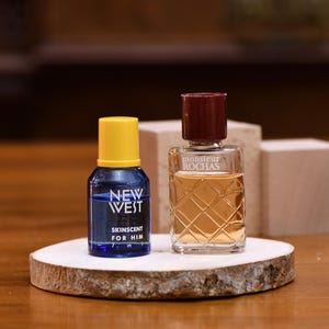 K&ouml;nnte beinhalten: Zwei kleine Parf&uuml;mflaschen auf einer Holzscheibe. Eine ist blau mit gelbem Deckel und tr&auml;gt die Aufschrift "NEW WEST SKINSENT FOR HIM". Die andere ist klar mit burgunderrotem Deckel und tr&auml;gt die Aufschrift "monsieur ROCHAS".