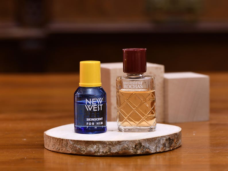 K&ouml;nnte beinhalten: Zwei kleine Parf&uuml;mflaschen auf einer Holzscheibe. Eine ist blau mit gelbem Deckel und tr&auml;gt die Aufschrift "NEW WEST SKINSENT FOR HIM". Die andere ist klar mit burgunderrotem Deckel und tr&auml;gt die Aufschrift "monsieur ROCHAS".