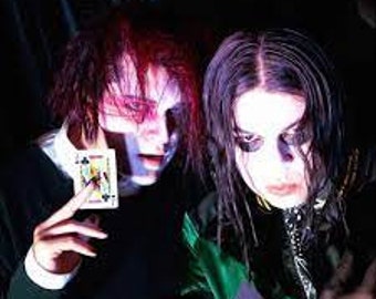 May include: Two people wearing dark clothing and white face paint, one holding a playing card. They are both looking at the camera.