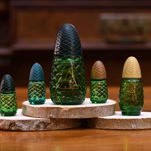 K&ouml;nnte beinhalten: Eine Sammlung gr&uuml;ner Glasparf&uuml;mflaschen in Ananasform mit strukturierten Oberfl&auml;chen und dunkelgr&uuml;nen, teal- und beigefarbenen Deckeln. Die Flaschen werden auf Holzscheiben pr&auml;sentiert und zeigen ein einzigartiges und dekoratives Design.