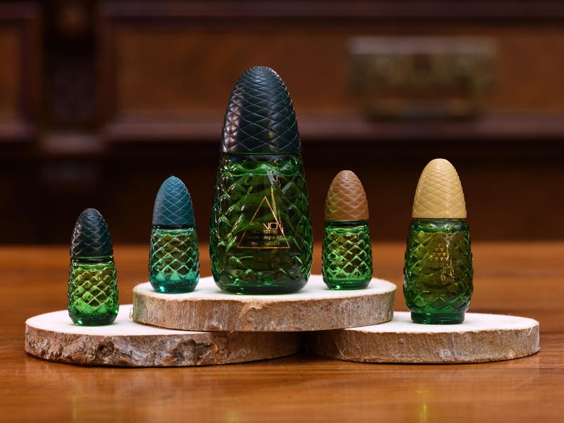 K&ouml;nnte beinhalten: Eine Sammlung gr&uuml;ner Glasparf&uuml;mflaschen in Ananasform mit strukturierten Oberfl&auml;chen und dunkelgr&uuml;nen, teal- und beigefarbenen Deckeln. Die Flaschen werden auf Holzscheiben pr&auml;sentiert und zeigen ein einzigartiges und dekoratives Design.