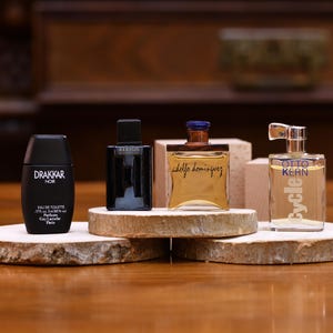 K&ouml;nnte beinhalten: Vier Parf&uuml;mflaschen in verschiedenen Formen und Gr&ouml;&szlig;en werden auf Holzplattformen pr&auml;sentiert. Die Flaschen umfassen eine schwarze Drakkar Noir-Flasche, eine schwarze Flasche mit dem Text "Keryus Givenchy", eine goldene Flasche mit dem Text "Adolfo Dominguez" und eine transparente Flasche mit dem Text "Otto Kern Cycle".