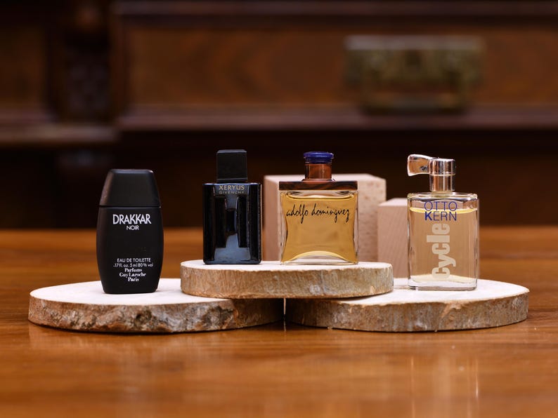 K&ouml;nnte beinhalten: Vier Parf&uuml;mflaschen in verschiedenen Formen und Gr&ouml;&szlig;en werden auf Holzplattformen pr&auml;sentiert. Die Flaschen umfassen eine schwarze Drakkar Noir-Flasche, eine schwarze Flasche mit dem Text "Keryus Givenchy", eine goldene Flasche mit dem Text "Adolfo Dominguez" und eine transparente Flasche mit dem Text "Otto Kern Cycle".