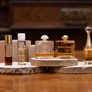 K&ouml;nnte beinhalten: Eine Auswahl an Parf&uuml;mflaschen, darunter eine hohe, schlanke Flasche mit goldenem Verschluss und eine transparente Flasche mit wei&szlig;em Verschluss. Die Flaschen werden auf Holzscheiben pr&auml;sentiert, die ihre verschiedenen Formen und Gr&ouml;&szlig;en zeigen. Der Hintergrund ist eine warme, verschwommene Umgebung.