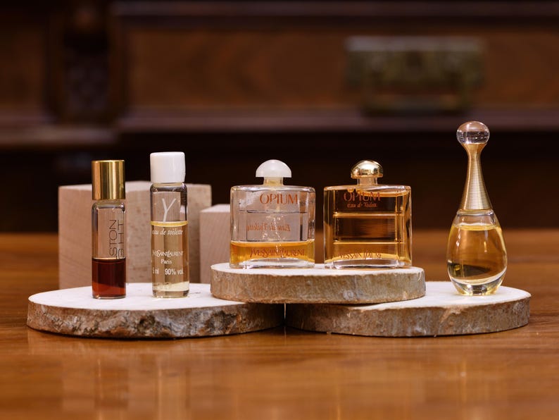 K&ouml;nnte beinhalten: Eine Auswahl an Parf&uuml;mflaschen, darunter eine hohe, schlanke Flasche mit goldenem Verschluss und eine transparente Flasche mit wei&szlig;em Verschluss. Die Flaschen werden auf Holzscheiben pr&auml;sentiert, die ihre verschiedenen Formen und Gr&ouml;&szlig;en zeigen. Der Hintergrund ist eine warme, verschwommene Umgebung.