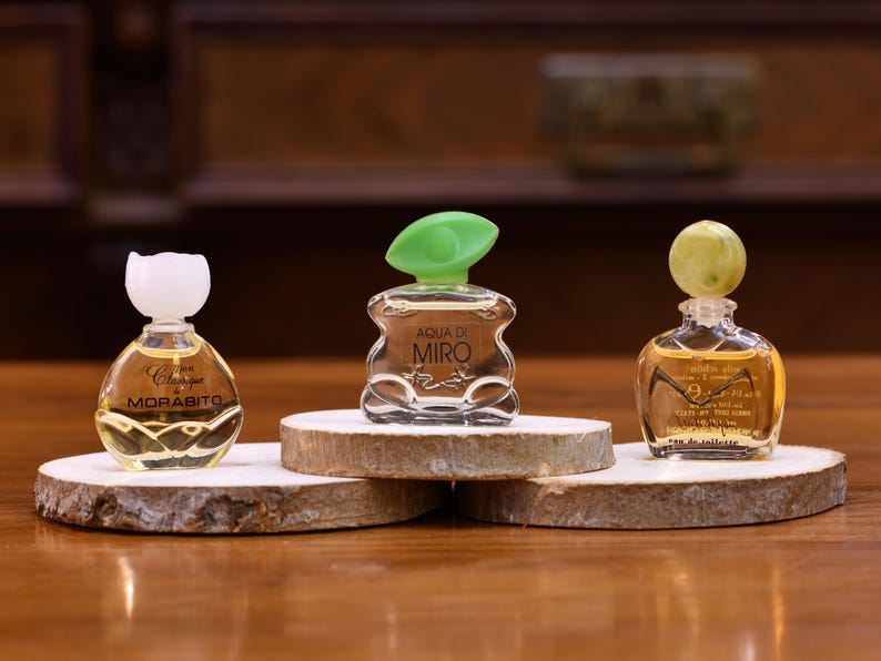K&ouml;nnte beinhalten: Drei Miniatur-Parf&uuml;mflaschen mit dekorativen Verschl&uuml;ssen werden auf Holzscheiben pr&auml;sentiert. Die Flaschen sind klar, mit einer goldenen Fl&uuml;ssigkeit gef&uuml;llt und mit Etiketten versehen. Die Verschl&uuml;sse sind wei&szlig;, gr&uuml;n und gelb. Die Flaschen sind auf einer Holzoberfl&auml;che angeordnet.
