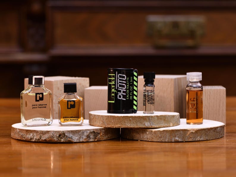 K&ouml;nnte beinhalten: Eine Auswahl an Parf&uuml;mflaschen und -fl&auml;schchen, darunter die Marken Paco Rabanne und Lagerfeld. Die Flaschen werden auf Holzplattformen und -bl&ouml;cken pr&auml;sentiert, wobei die Lagerfeld-Flasche mit "PHOTO" beschriftet ist. Die D&uuml;fte sind in verschiedenen Gr&ouml;&szlig;en und Farben erh&auml;ltlich.