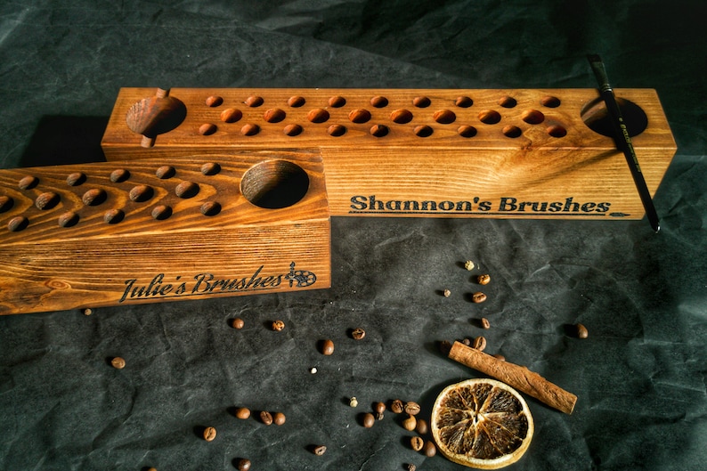 Pu&ograve; includere: Due portapennelli in legno con finitura rustica. Uno &egrave; etichettato "Julie's Brushes" e l'altro "Shannon's Brushes". Ognuno ha pi&ugrave; fori per pennelli e un foro pi&ugrave; grande. Un pennello poggia su uno. Chicchi di caff&egrave; e una fetta d'arancia secca sono sparsi.
