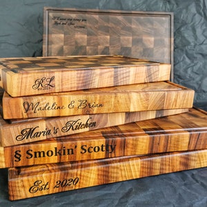 Puede incluir: Una pila de cinco tablas de cortar de madera con diferentes grabados personalizados. La tabla superior tiene las iniciales "RL" y la fecha "est. 2021". La segunda tabla tiene los nombres "Madeline & Brian". La tercera tabla tiene el texto "Maria's Kitchen". La cuarta tabla tiene el texto "&sect; Smokin' Scotty". La tabla inferior tiene el texto "Est. 2020".