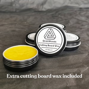 Puede incluir: Un primer plano de un peque&ntilde;o tarro de cera para tabla de cortar totalmente natural. El tarro es negro con una etiqueta blanca que dice "GrainWoods Cutting Board Wax All-Natural". Hay varios tarros de cera en la imagen.