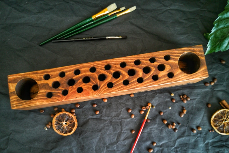 Pu&ograve; includere: Un portapennelli in legno con numerosi fori e due grandi aperture circolari. Diversi pennelli con manici verdi e setole dorate sono appoggiati su una superficie scura. Chicchi di caff&egrave; e una fetta d'arancia essiccata sono sparsi.