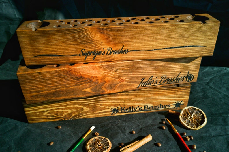 Pu&ograve; includere: Tre portapennelli in legno impilati. Il portapennelli superiore ha diversi piccoli fori e un foro pi&ugrave; grande. Il testo "Supriya's Brushes" &egrave; inciso sul portapennelli superiore. I testi "Julie's Brushes" e "Kelly's Brushes" sono incisi sugli altri.
