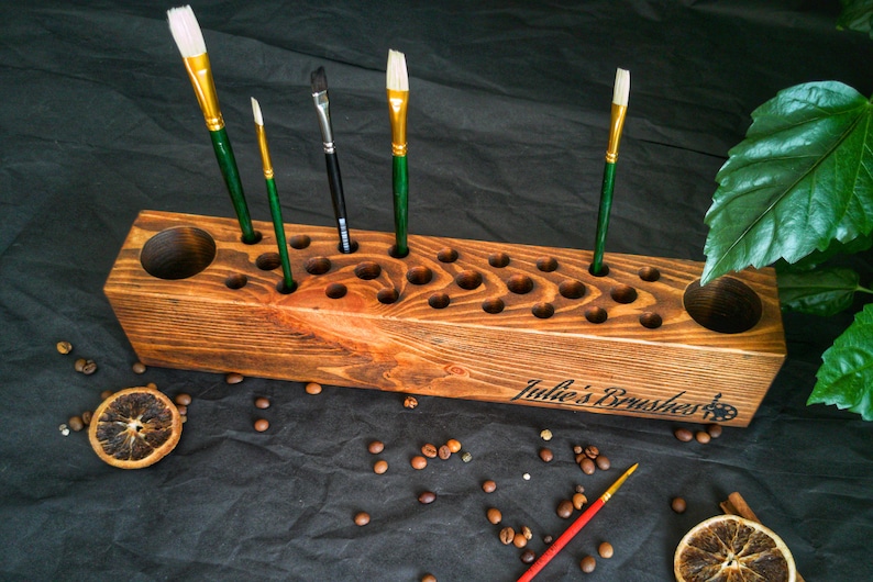 Pu&ograve; includere: Un portapennelli in legno marrone con fori, che contiene pennelli con manici verdi e setole bianche. Il supporto ha l'incisione "Julie's Brushes". Chicchi di caff&egrave; e fette d'arancia essiccate sono sparsi intorno.