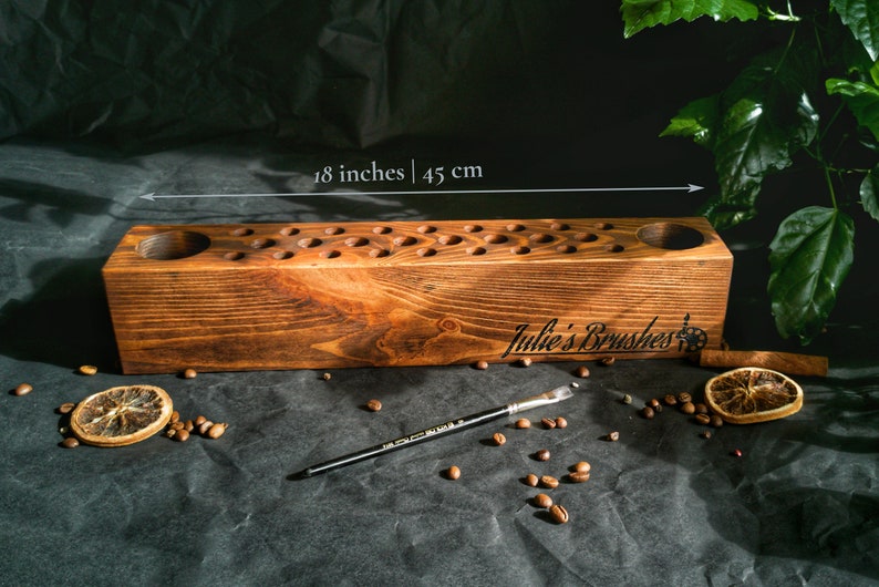 Pu&ograve; includere: Un portapennelli in legno tinto, con numerosi piccoli fori e due aperture circolari pi&ugrave; grandi. Il supporto &egrave; lungo 45 cm e presenta la scritta "Julie's Brushes". Sono visibili anche un pennello, chicchi di caff&egrave; e fette d'arancia essiccate.