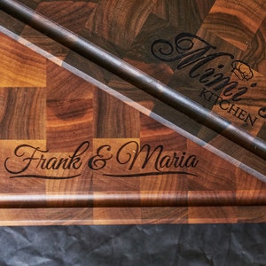 Puede incluir: Dos tablas de cortar de madera con texto grabado. La tabla superior tiene "Mimi's Kitchen" grabado. La tabla inferior tiene "Frank & Maria" grabado.
